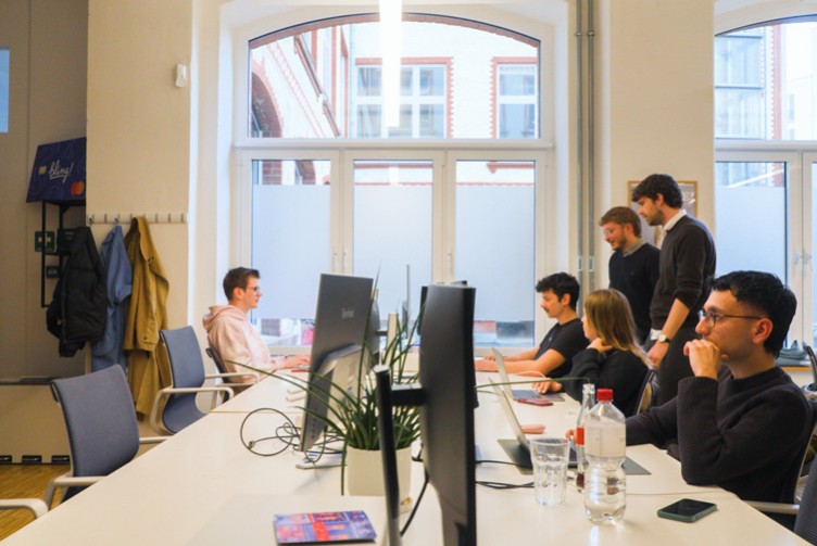 Mehrere Personen arbeiten in einem hellen Büro mit großen Fenstern an Computern und Laptops