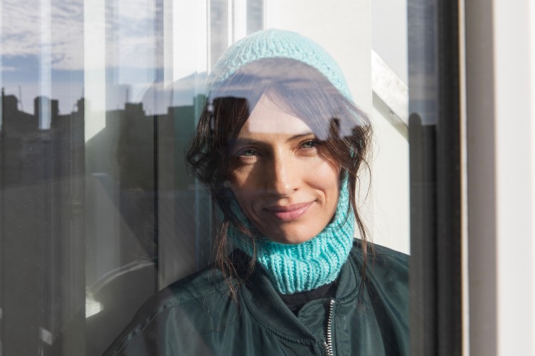 Weibliches Model mit braunen Haaren und türkisfarbener gestrickter Mütze steht vor einer Fensterscheibe.