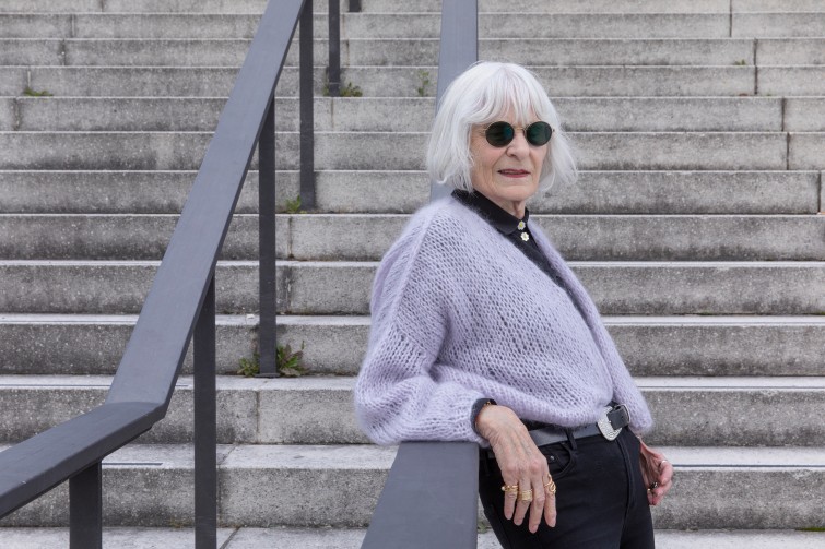 Model mit weißen Haaren und Sonnenbrille steht an einem Metallgeländer angelehnt und trägt eine selbstgestrickte Jacke in Flieder. 