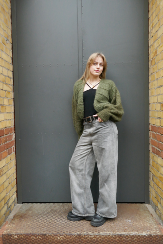 Eine junge Frau lehnt an einer Metalltür und trägt eine Strickjacke in Oliv.
