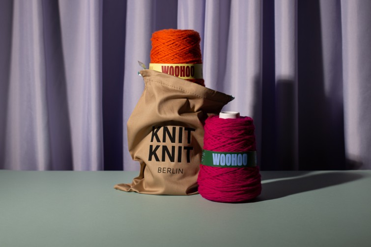 Zwei Wollknäul drapiert auf einer Unterlage, eines in einer Tasche mit der Aufschrift KnitKnit.