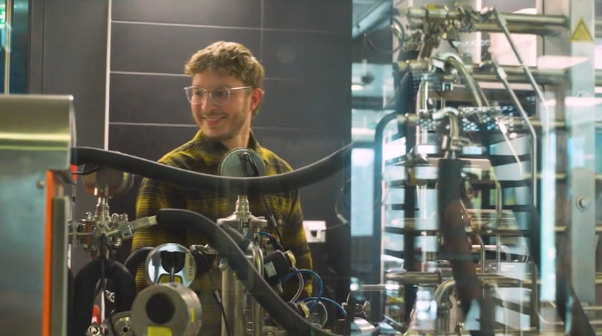 Robert (von der IBB) steht lächelnd inmitten einer industriellen Produktionsanlage