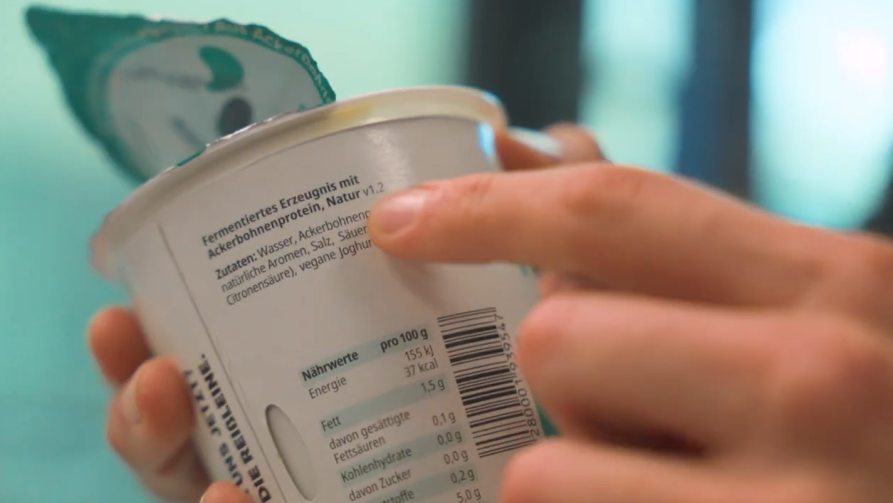 Hand hält einen geöffneten vly-Becher, auf dem die Zutatenliste und Nährwertangaben zu lesen sind