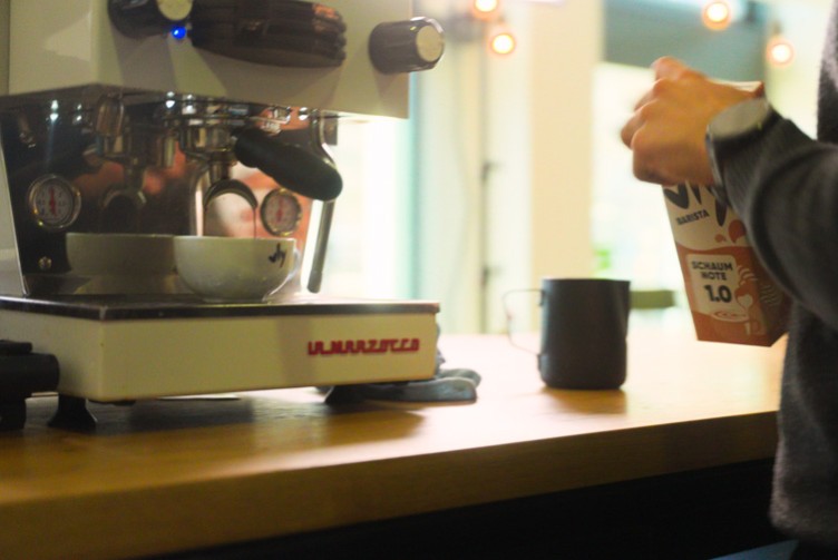 Hand gießt vly Barista-Drink in ein Milchkännchen neben einer Espressomaschine